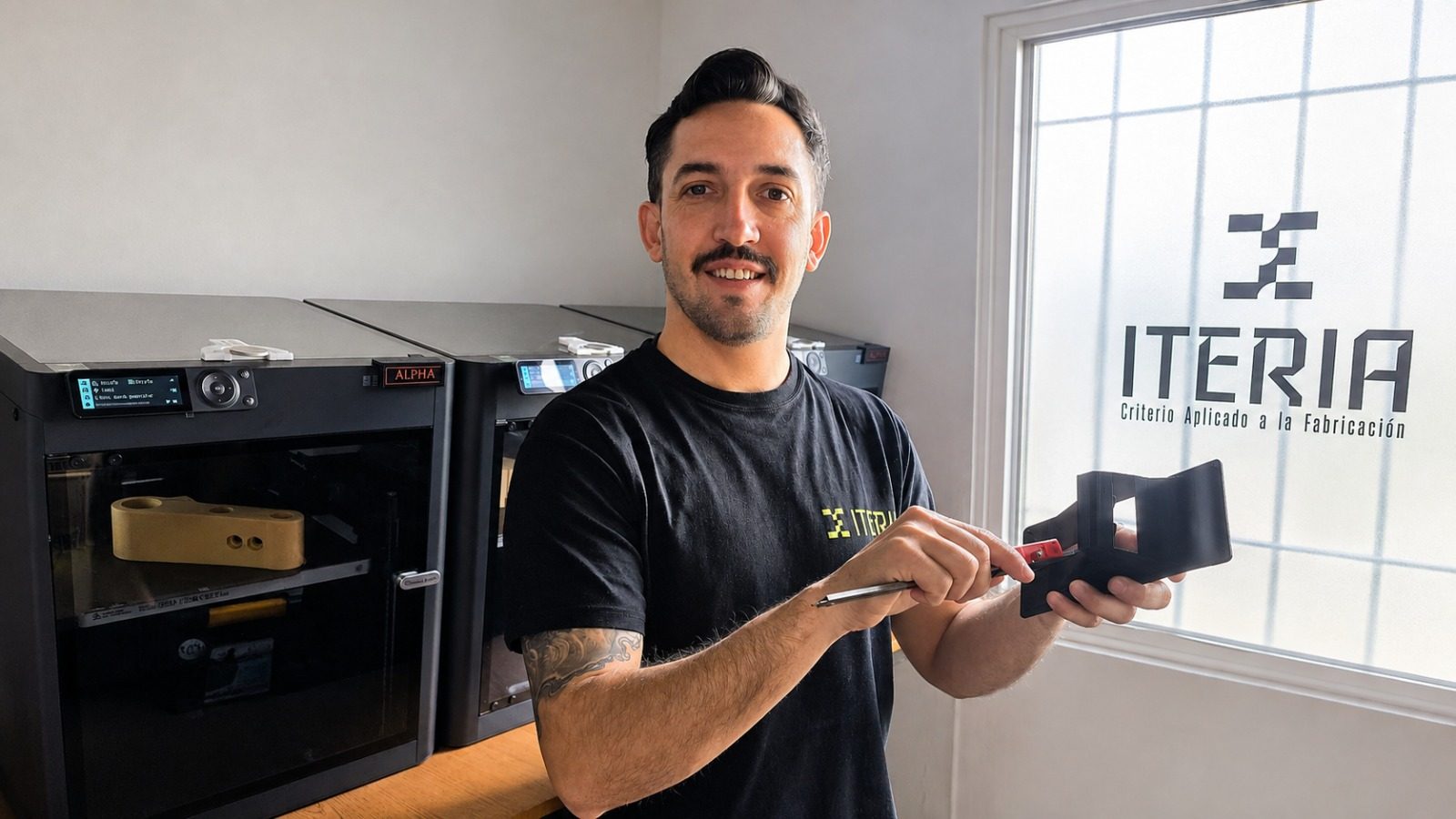 Facundo Vicente en el taller de Iteria, sosteniendo una pieza técnica impresa en negro junto a dos impresoras 3D, con el logo de Iteria en la ventana