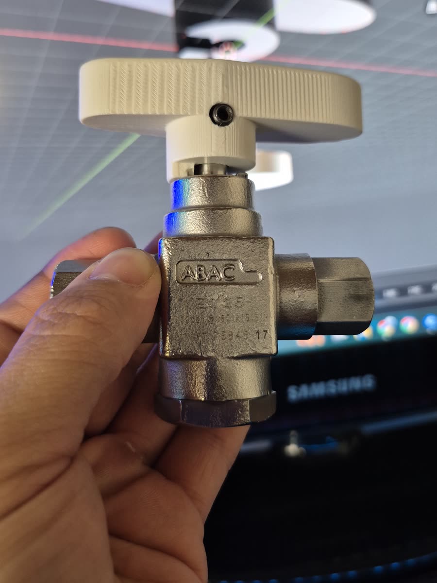 Válvula de bronce con la manija de accionamiento reimpresa ya montada, sostenida en la mano frente al monitor donde se ve el modelo CAD de la misma pieza